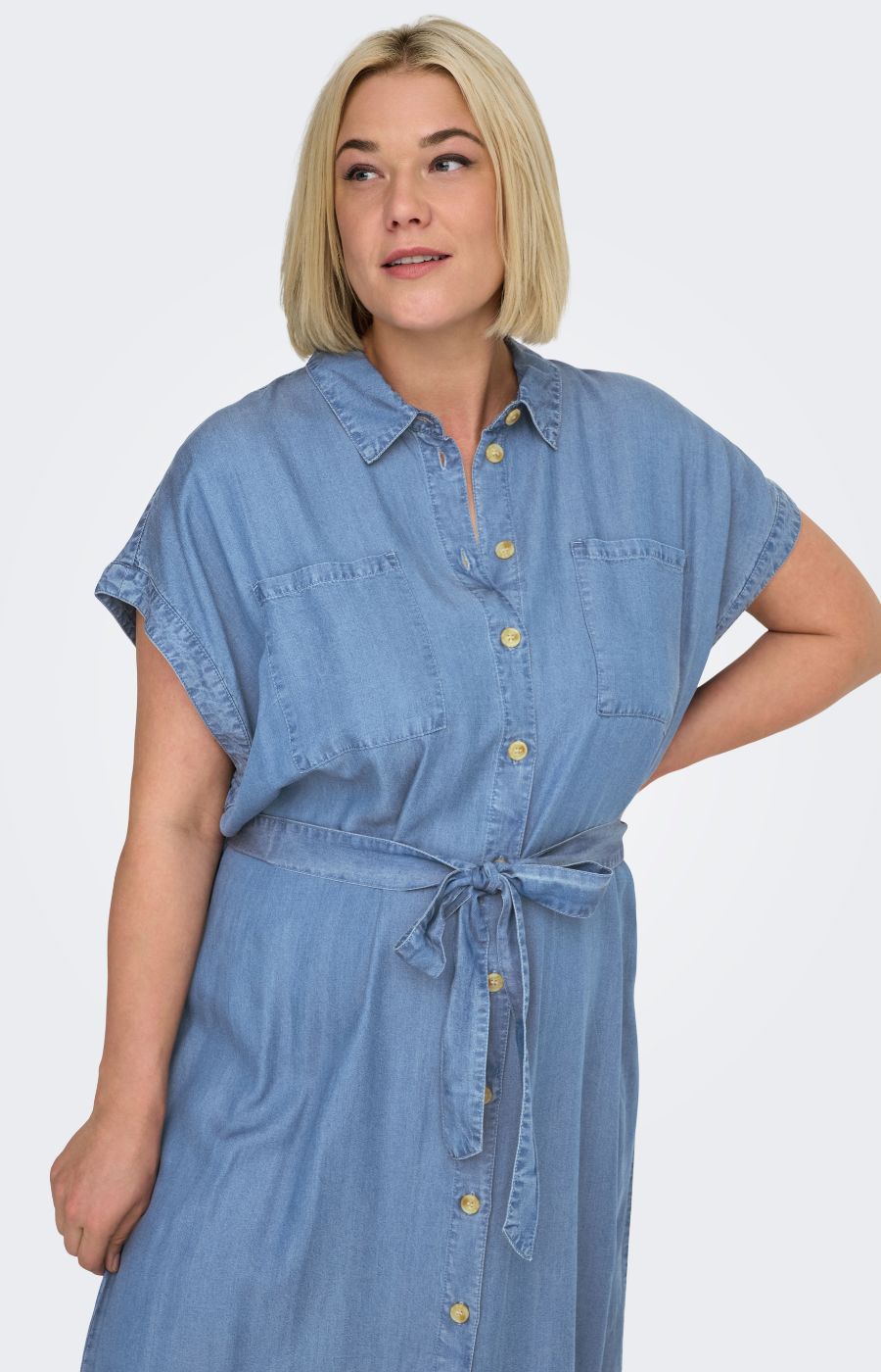 Woman wearing the Only Carmakoma Pema Denim Dress, a button up dress with a collared neck and breast pockets and a tie belt against a white background