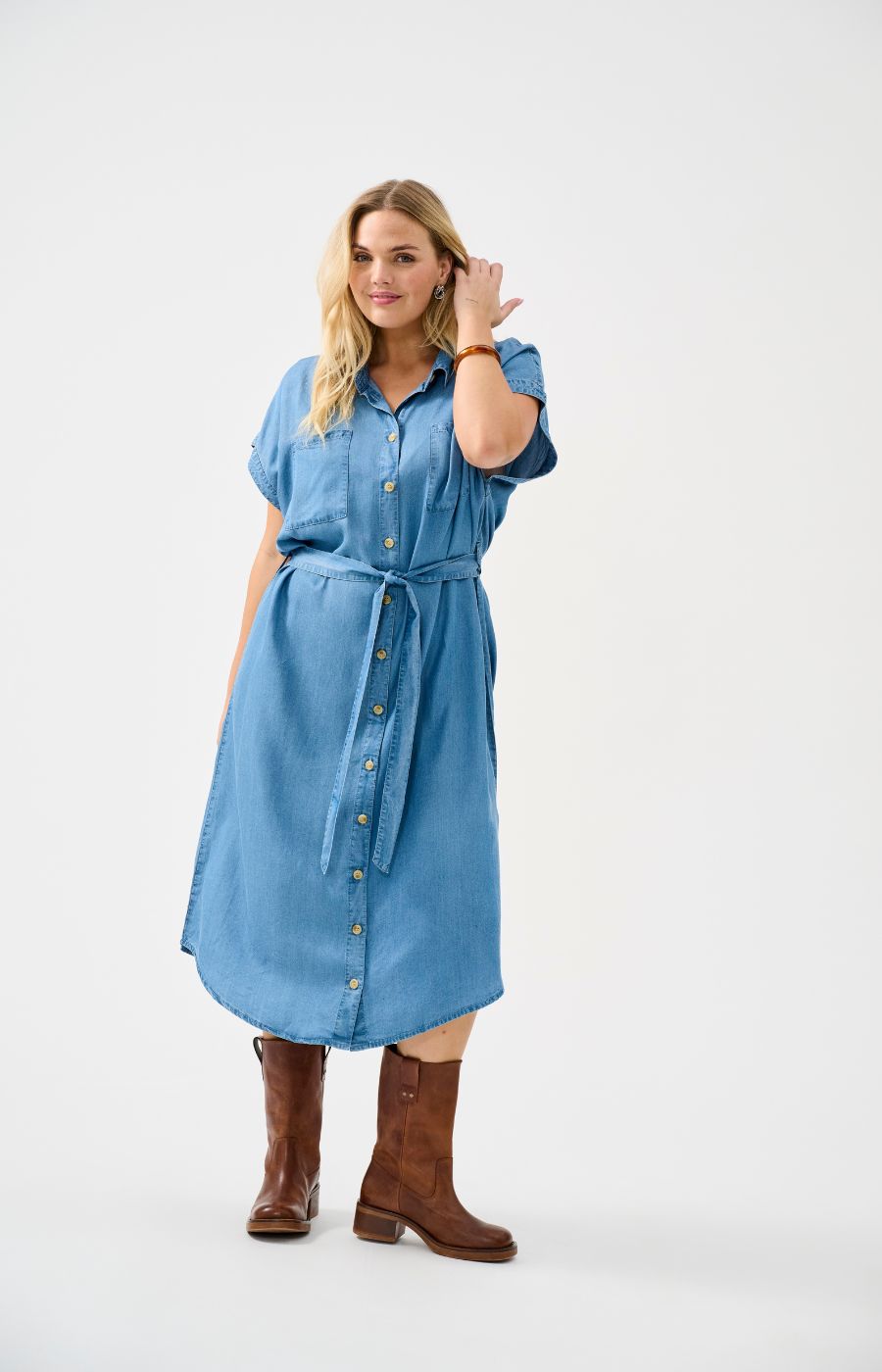 Woman wearing the Only Carmakoma Pema Denim Dress, a blue denim dress with brown knee-high boots on a white background