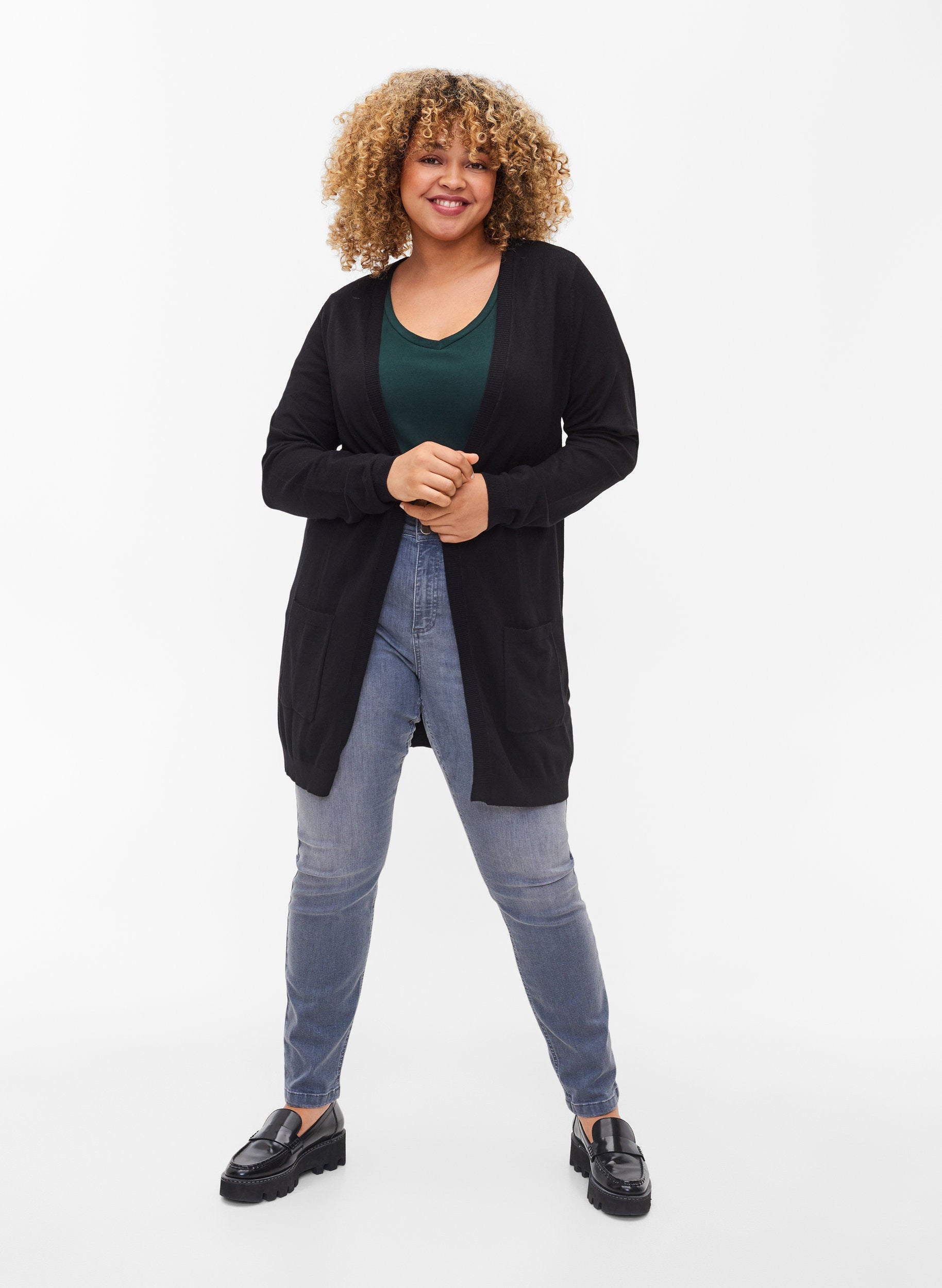 Woman wearing a black long line, open cardigan with long sleeves, green top, and blue jeans on a white background