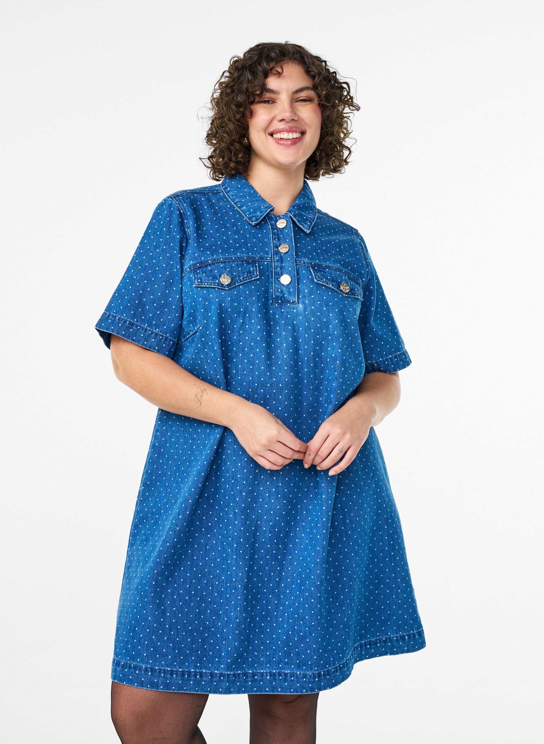 Woman wearing the Zizzi Misha Denim Dress, A blue polka dot dress with a shirt collar, short button placket, knee length and with short sleeves  on a white background